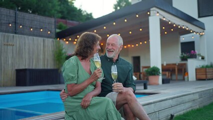 Senior man with his wife celebrating birthday and toasting with wine near backyard pool.