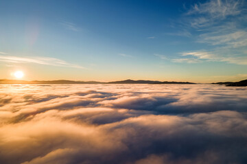 Aerial view of vibrant yellow sunrise over white dense clouds and distant mountains on horizon