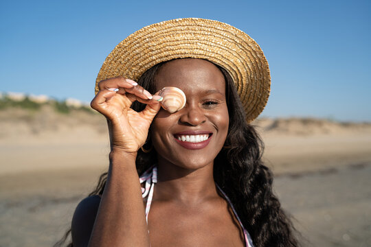 Cheerful black woman covering eye with seashell
