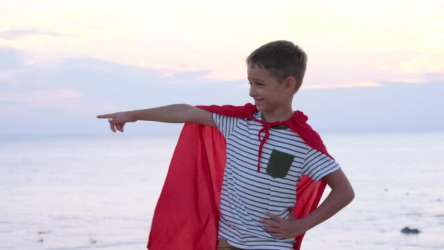 A happy kid is playing super hero against the background of the sky and the sea, pointing into the distance.The concept of power, happy childhood, childhood dreams. forward