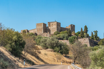 Road and hillside with dry herbs and trees and Monsaraz fortress castle in the background, Alentejo PORTUGAL