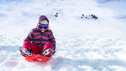 Fototapeta na wymiar Happy little girl going down the hill on sleigh in the mountain resort
