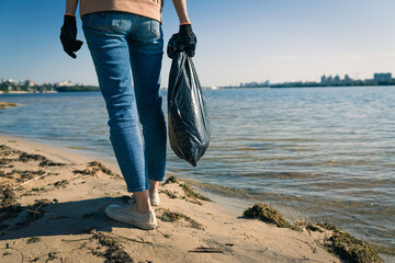 Woman with black plastic bag going on sand beach for collecting garbage. Earth Day or Environment Day