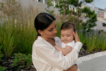 Close up outside photo of young adorable woman with dark hair holding little son during waking in summer city 