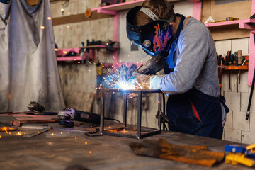 Metalworker in safety mask and gloves using welding torch to join sides of steel cube standing at...