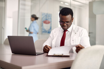Black general practitioner analyzing medical reports at doctor's office.