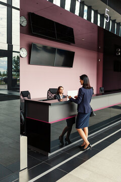 Middle aged businesswoman talking with young female receptionist at counter while checking in at hotel or office reception, wide shot side view