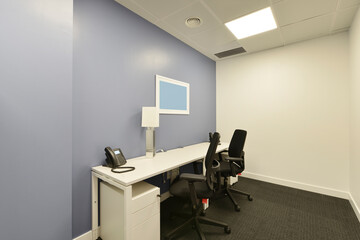 Small office cubicle with a white table, two chests of drawers, two twin black swivel chairs and dark gray carpet floors