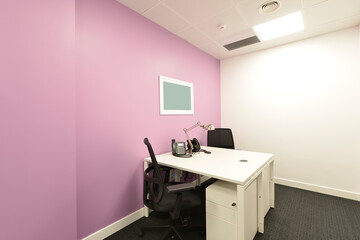 Small office cubicle with a double white desk, matching chest of drawers, black swivel chairs and switchboard phones
