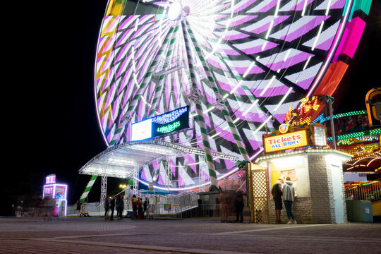Tourists purchase tickets and enjoy entertainment at a night time scene lit by the glowing midway lights of a large ferris wheel operated at a local carnival.