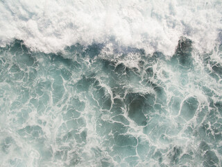 wide overhead view of violently crashing waves in a rough surf of emerald blue waters, creating thick white sea foam on top of the ocean as the strong undertow current retreats