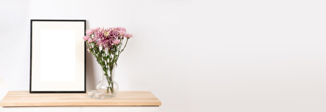 A mock-up of a black photo frame on wooden table, a transparent vase with pink summer flowers, banner size