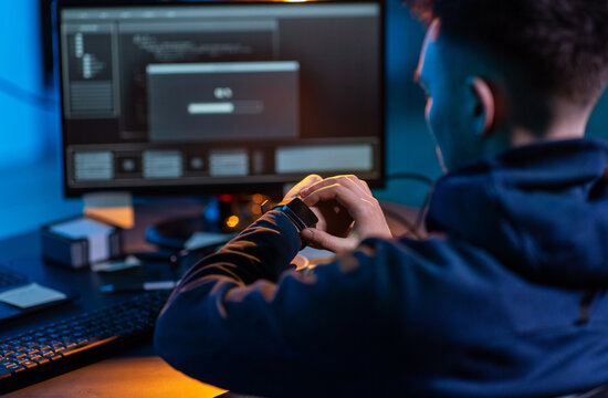 cybercrime, hacking and technology concept - close up of male hacker in dark room with smart watch and computer carries out cyber attack in dark room