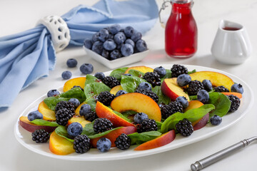 Summer fruit salad with mixed berries, peach slices and fresh green on white plate with fresh blueberry and dressing on background.
