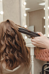 Fototapeta premium girl in beauty salon is doing her hair. women's hair styling on flat iron. blog aesthetics, beauty, hair stylists. hair curling curling iron, space for text.