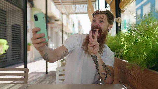 Young redhead man making selfie by the smartphone sitting on table at coffee shop terrace