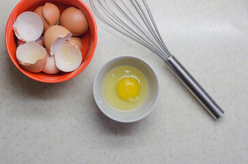 The shell of broken eggs, a broken egg in a bowl and a whisk on the table