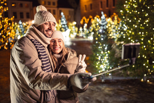 winter holidays and people concept - happy smiling couple taking picture with smartphone on selfie stick over christmas tree lights in evening city