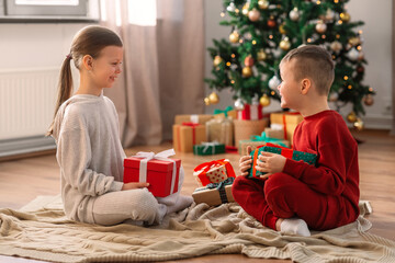 Obraz na płótnie Canvas christmas, winter holidays and childhood concept - happy girl and boy in pajamas with gifts sitting on floor in front of each other at home
