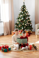 Obraz na płótnie Canvas christmas, winter holidays and childhood concept - happy girl and boy in pajamas reading book together sitting on floor in front of each other at home