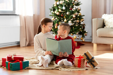 christmas, winter holidays and childhood concept - happy girl and boy in pajamas reading book together sitting on floor in front of each other at home