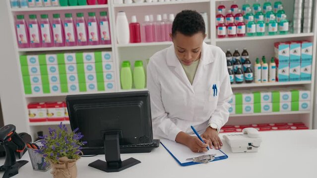 African american woman pharmacist writing on document using computer at pharmacy