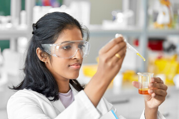 science research, work and people concept - female scientist in goggles with dropper and chemical or test sample in beaker working in laboratory