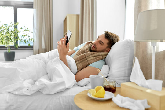 health, illness and people concept - sick man with smartphone in bed at home