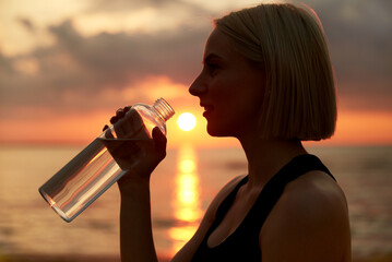 fitness, sport, and healthy lifestyle concept - close up of woman drinking water from bottle on beach over sunset
