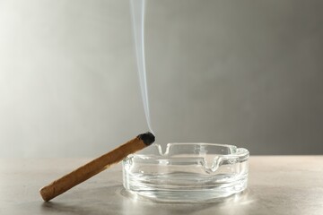 Smoldering cigar near glass ashtray on light grey table, space for text