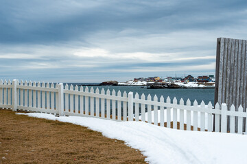 A grey wooden building with a white picket fence attached and enclosing the boundary of a yard overlooking the blue ocean with a cloudy sky. There's white snow on the ground and land in the background - Powered by Adobe