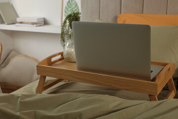 Wooden tray with modern laptop and burning candle on bed indoors