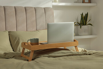 Wooden tray with modern laptop and cup of aromatic drink on bed indoors