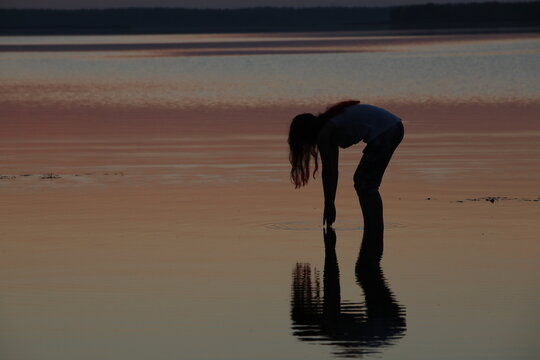Silhouette of a girl in summer at sunset standing in the river bending down with her hands outstretched to the water