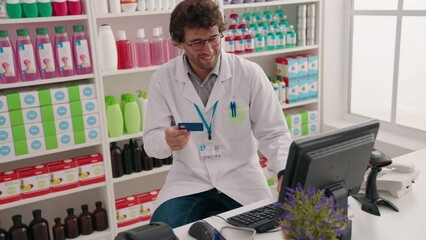 Young hispanic man pharmacist using computer holding credit card at pharmacy - Powered by Adobe