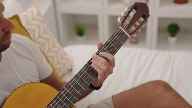 Young hispanic man playing classical guitar sitting on bed at bedroom