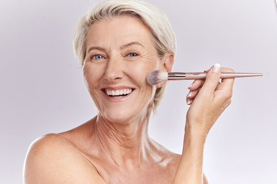 Beauty, makeup and cosmetics of senior woman with a make up brush applying foundation, concealer or cosmetic product with a smile portrait. Happy blonde lady showing skincare for reducing wrinkles