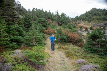 A senior aged man wearing a blue coat and hiking pants uses a handheld GPS to navigate a hiking trail. There are trees, large rocks, and shrubs among a worn path. The sky is cloudy white with blue. 