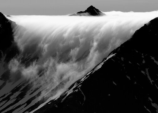 Black and white image of a dramatic mountain landscape with clouds flowing over rugged peaks.