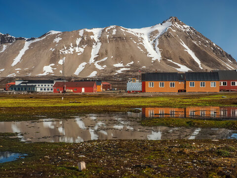 Colorful wooden houses in a remote Arctic settlement with snow-capped mountains and reflections in the foreground. This photograph captures a typical polar village scene during the summer thaw, featur