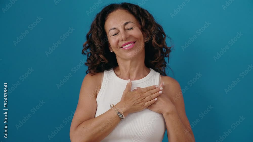 Poster middle age woman smiling confident standing with hands on heart over isolated blue background