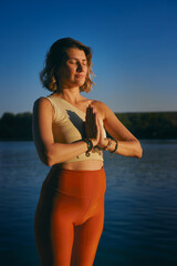 A yogi woman is doing sun salutation near the water on a dock at sunset.