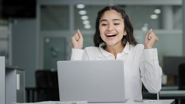 Young woman manager sitting in office looking at laptop screen receiving notification reads good news rejoices in success celebrating victory feels good luck receives promotion at work gets job bonus