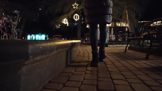 Woman's feet walk through city, through park with lights at night. It's cold outside, she walks around in her boots and coat. Christmas or holiday. Camera follows her on back. close-up of legs.
