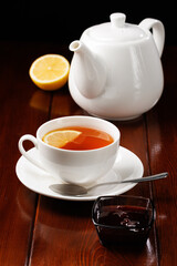 Tea with lemon in a porcelain cup next to a white teapot on a wooden table background. Angle view. Selective focus. Vertical frame orientation.