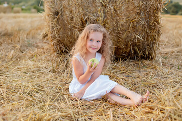 a little curly-haired girl is sitting by a haystack in a field with a green apple in her hands