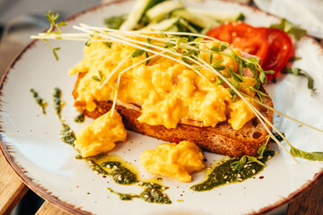 A slice of toast bread with a soft egg on it and some greens. Some slices of tomatoes and cucumbers next to it, and some pesto as decoration on a plate