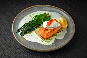 Grilled salmon fillet with zucchini, spinach, cream cheese, garnished with red caviar with cream sauce in a ceramic plate on a dark textured background. Restaurant menu Isolated on black