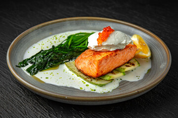 Grilled salmon fillet with zucchini, spinach, cream cheese, garnished with red caviar with cream sauce in a ceramic plate on a dark textured background. Restaurant menu Isolated on black