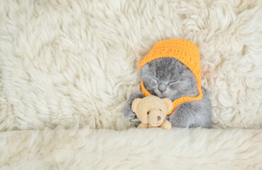 Cozy tiny kitten wearing tiny warm hat sleeps with favorite toy bear under warm blanket on a bed at...